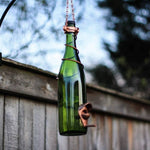 Green glass bottle bird feeder hanging against a wooden fence with blurred trees in the background
