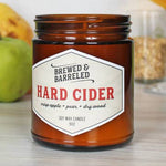 Candle labeled 'Brewed & Barreled Hard Cider' on a wooden surface with apples in the background.