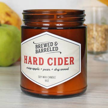Candle labeled 'Brewed & Barreled Hard Cider' on a wooden surface with apples in the background.