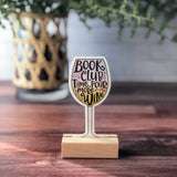 Sticker shaped like a wine glass with 'Book Club Time. Pour More Wine' text displayed on a wooden surface.